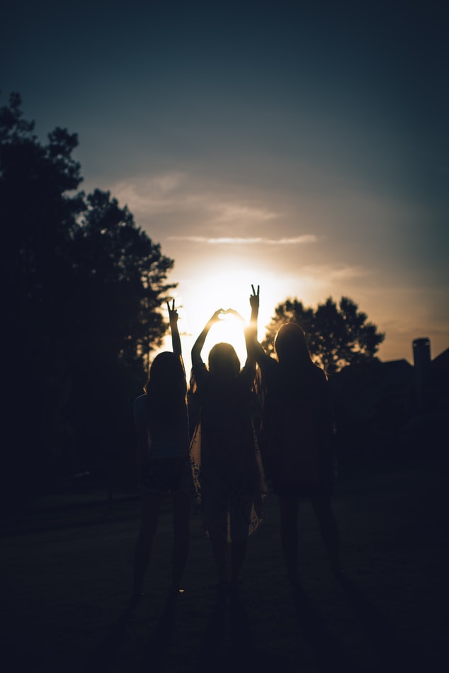 Silhouettes of 3 girls reaching up with peace signs in front of a sunset